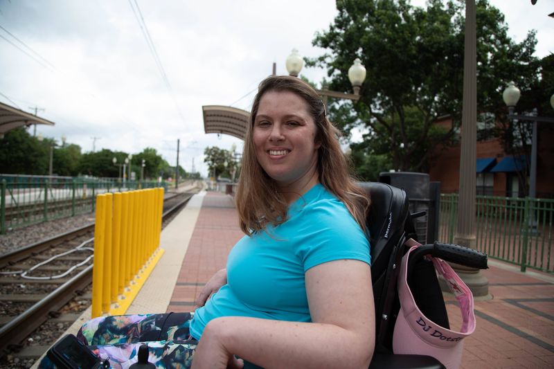 A smiling woman in a power wheelchair waits on a train platform.