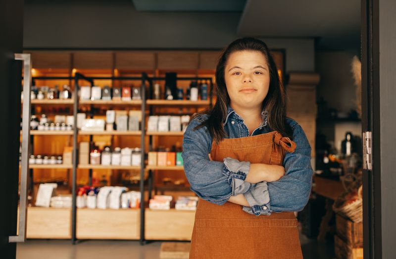 A young woman in an apron stands with her arms crossed in the shop where she works.